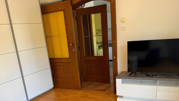 A room with a wooden floor, a white sofa, a TV on a stand, and a door leading to another room.