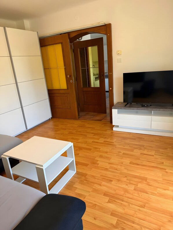 A room with a wooden floor, a white sofa, a TV on a stand, and a door leading to another room.