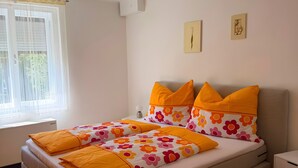 A bedroom with a bed, a window with blinds, a wall clock, and a small bedside table.