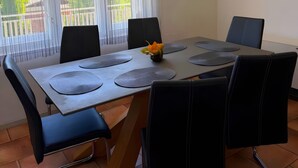 A dining area with a table set for four, surrounded by black chairs, a flower centerpiece, and a view of greenery through sheer curtains.
