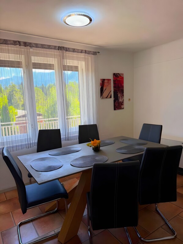 A dining area with a table set for four, surrounded by black chairs, a flower centerpiece, and a view of greenery through sheer curtains.