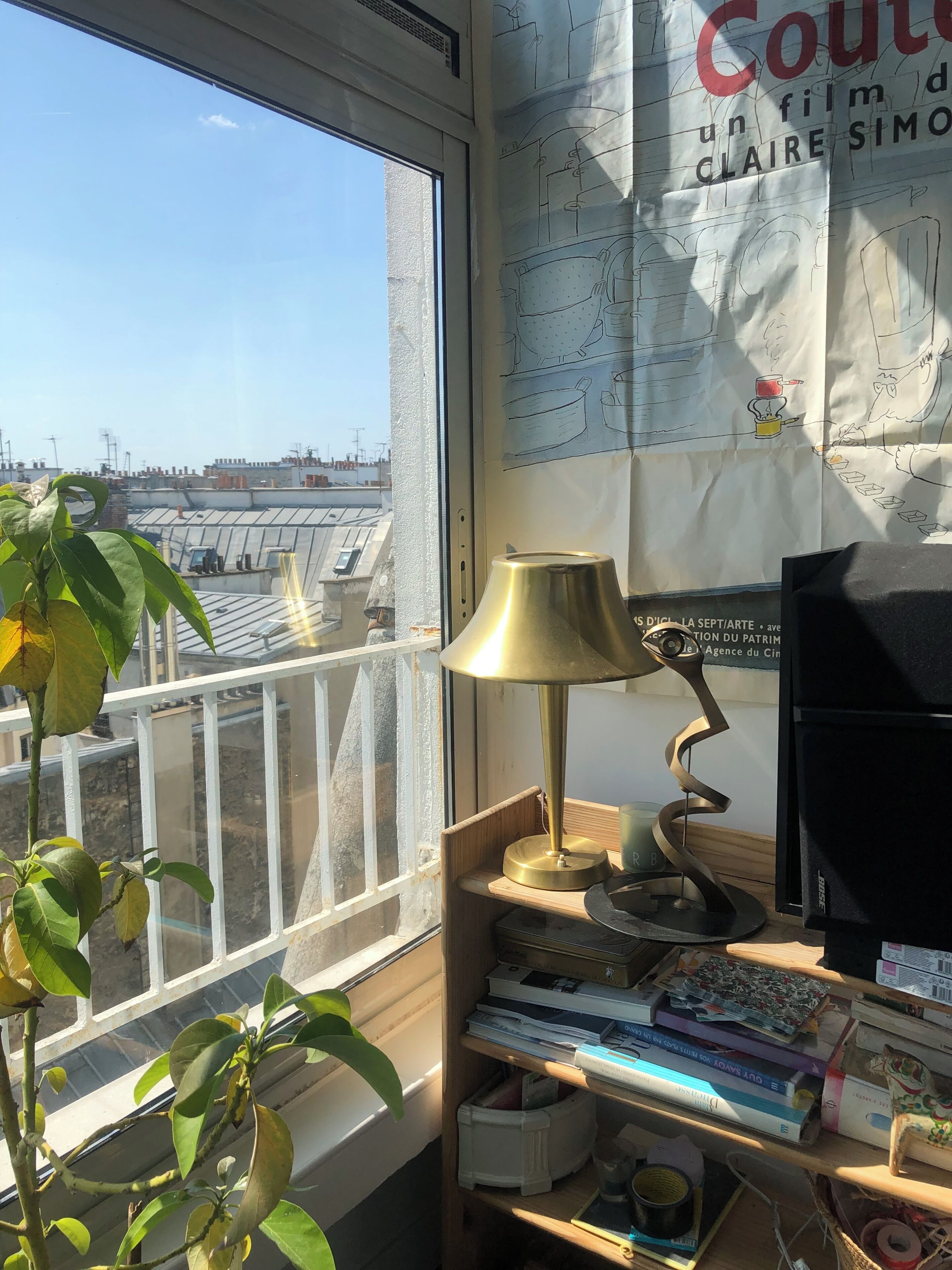 Mesa com luminária, livros e monitor de computador. Janela com vista para edifícios visível.