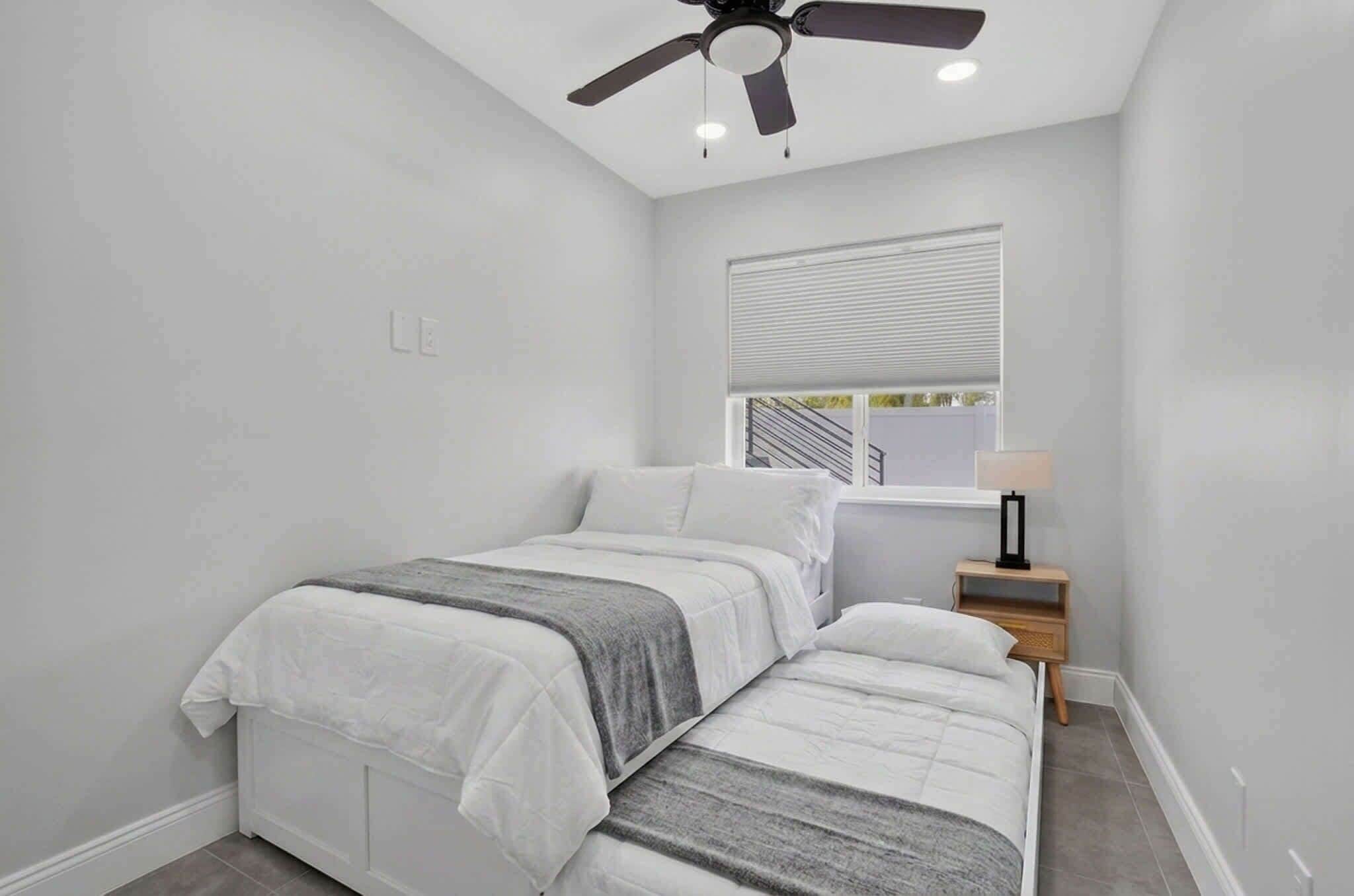 Ein Schlafzimmer mit einem Bett, einem Nachttisch, einem Deckenventilator und einem Fenster mit Jalousien.
