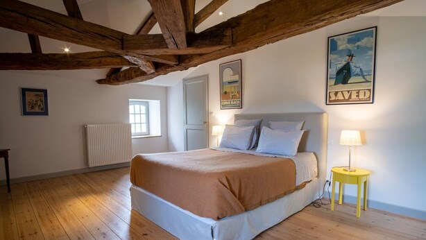 Quarto com teto de madeira, cama com manta marrom, abajur e um pôster na parede.