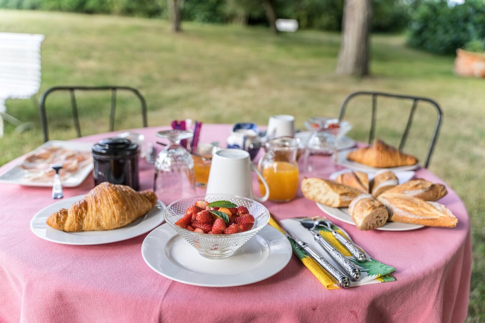 Un petit-déjeuner en plein air, avec des croissants, des fraises, du jus de fruits et du café, disposés sur une nappe rose.