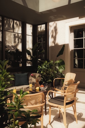 A sunlit room with a sofa, two wicker chairs, a glass table with drinks, and large windows.