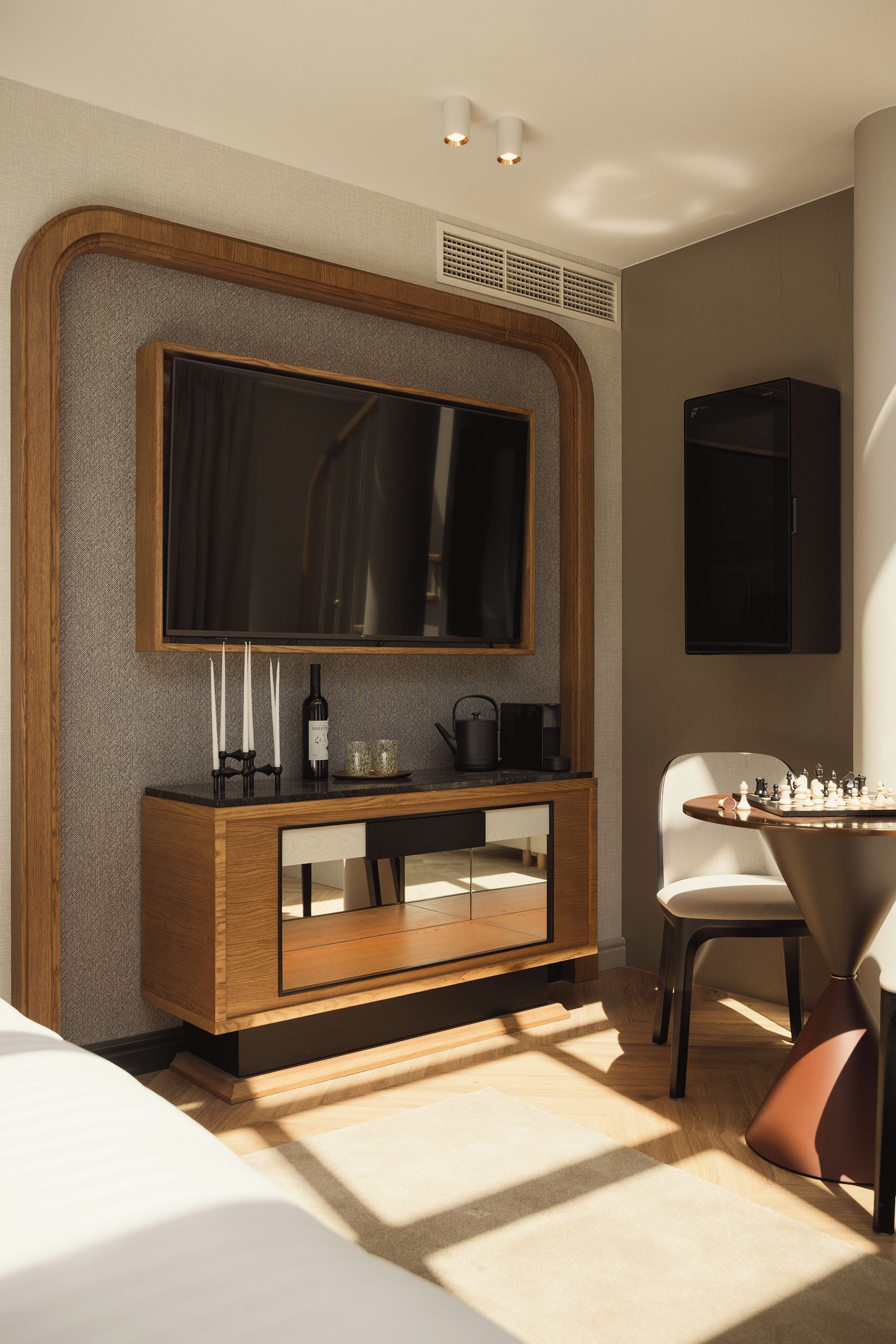Habitación de hotel moderna con un televisor de pantalla plana grande montado en una pared texturizada, una mesita de madera con una botella de vino y copas, una cafetera y un tablero de ajedrez sobre una mesa redonda.