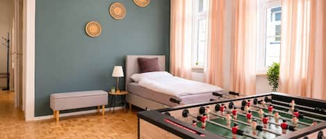 A room with a bed, a foosball table, a bench, and a small table with a lamp.