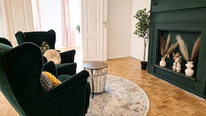 Ett rum med en grön fåtölj, ett litet runt bord, en mönstrad matta, en öppen spis med dekorativa föremål och en dörr med gardiner.
