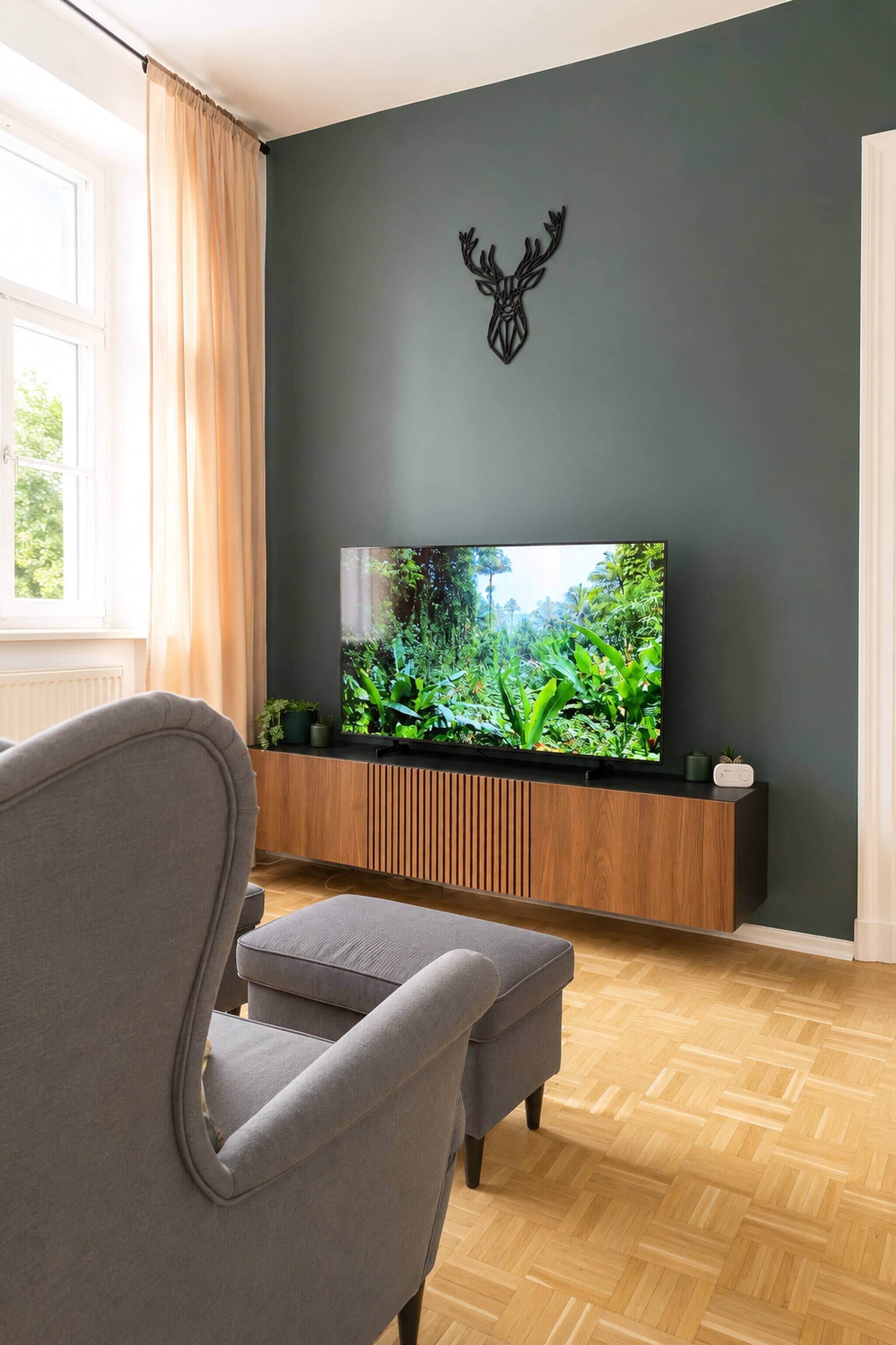 A modern living room with a flat-screen TV mounted on a wooden console, a grey armchair, and a decorative deer head on the wall.