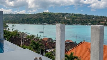 A view from a balcony overlooking a turquoise sea with boats, a hilly landscape, and a red-roofed building in the foreground.
