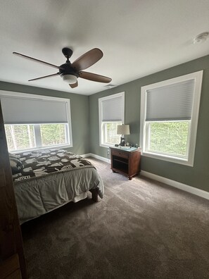 Quarto com cama, mesa de cabeceira, ventilador de teto e duas janelas com persianas.