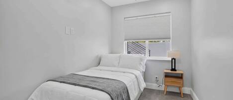 Een slaapkamer met een bed, een plafondventilator, een raam met jaloezieën, een nachtkastje met een lamp en een stopcontact.