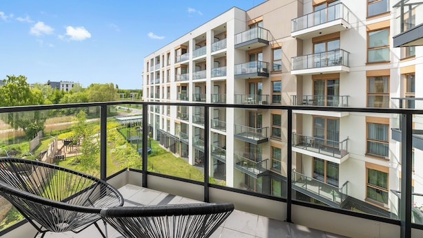 A modern apartment building with multiple balconies, a clear view of the surrounding greenery, and a balcony with a chair and umbrella.