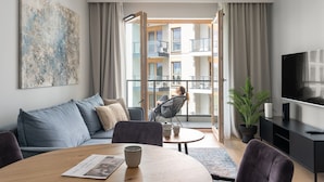 A modern living room with a sofa, a round table with a newspaper, a chair, a television, and a balcony with a view of a building.