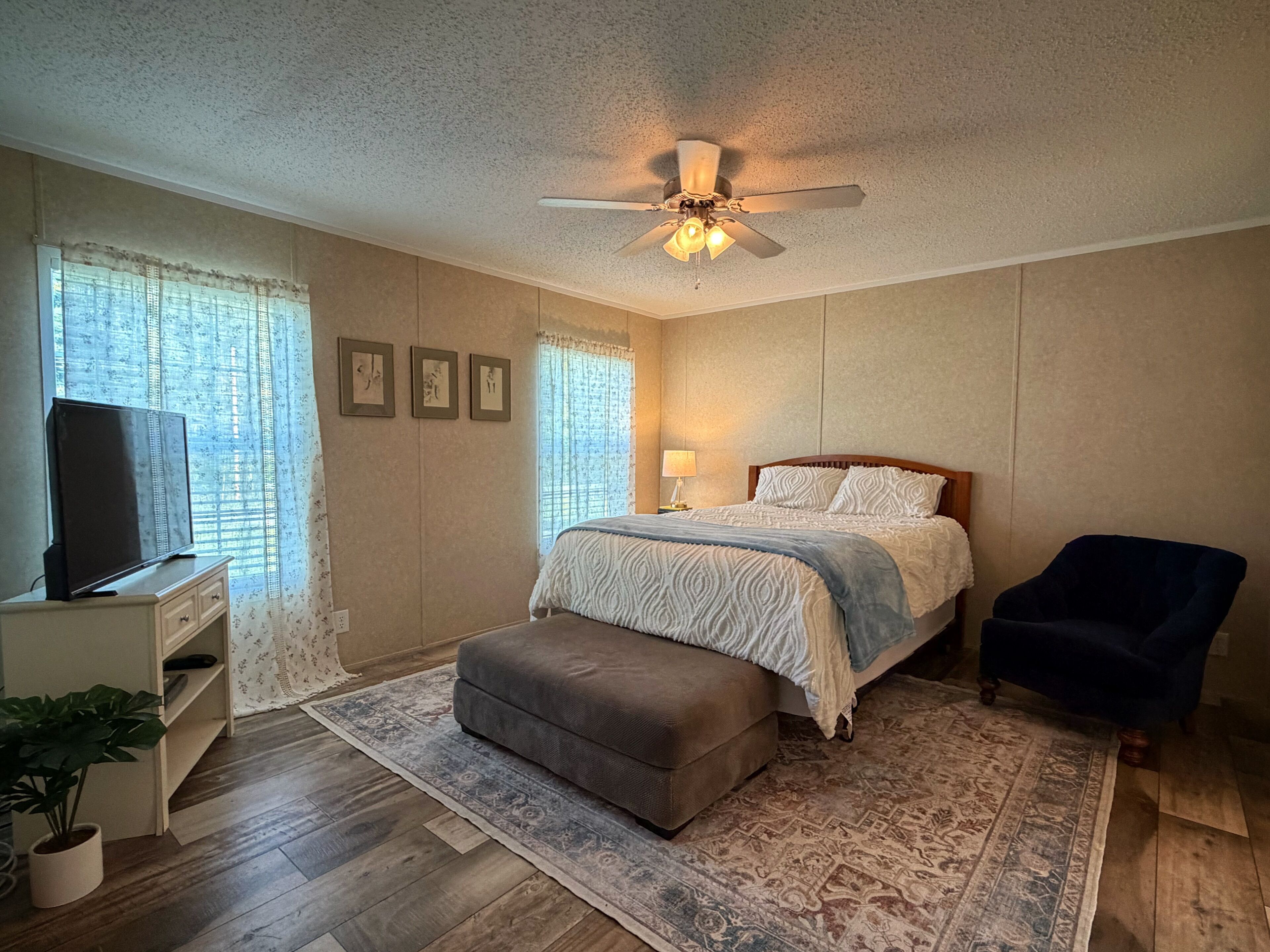 A bedroom with a bed, a chair, a television, a ceiling fan, and a window with curtains.