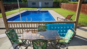 Un patio trasero con piscina, un porche cubierto con mesa y sillas, y una valla de madera.