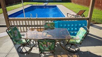 Un jardin avec une piscine, une terrasse couverte dotée d’une table et de chaises, et une clôture en bois.