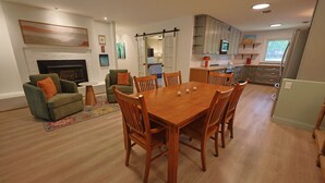 Uma sala de estar ampla com lareira, sala de jantar com mesa e cadeiras de madeira, e cozinha com armários em tons claros.