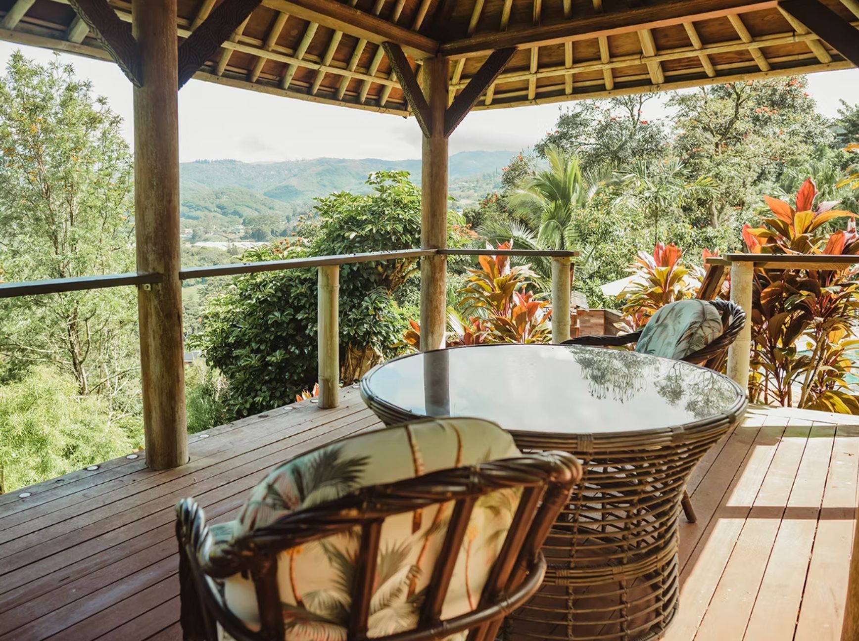 A wooden deck with a glass-top table and wicker chairs, overlooking a lush landscape with trees and hills.