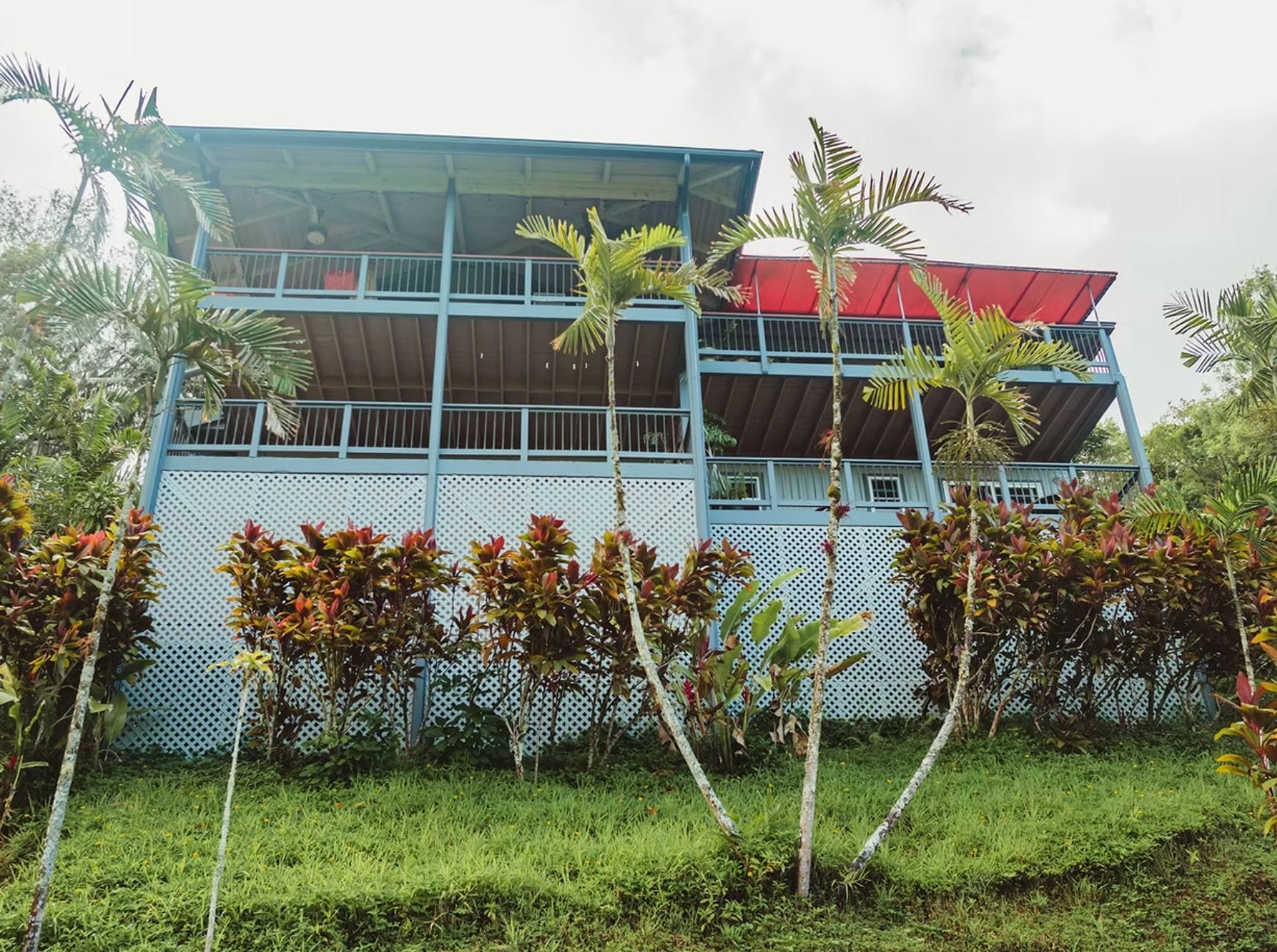 Un edificio de dos pisos con fachada azul y techo rojo, rodeado de abundante vegetación.