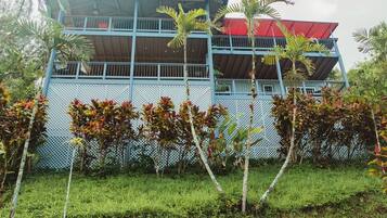Un edificio de dos pisos con fachada azul y techo rojo, rodeado de abundante vegetación.