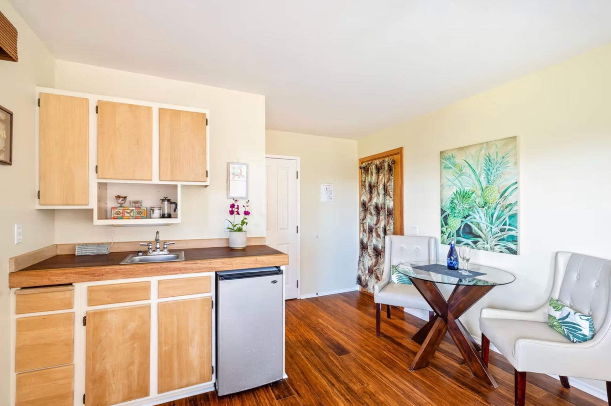 A compact kitchen with wooden cabinets, a built-in refrigerator, and a dining area with a glass table and white chairs.