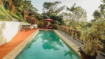 Una piscina con un paraguas rojo, sillas de descanso y un jardín con diversas plantas.