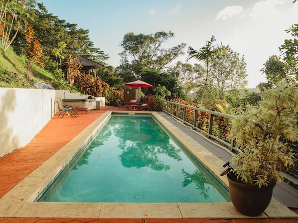 A swimming pool with a red umbrella, lounge chairs, and a garden area with various plants.
