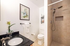 A bathroom with a sink, toilet, shower, and a framed picture on the wall. - Marjorie Kauai Inn (Lawai)