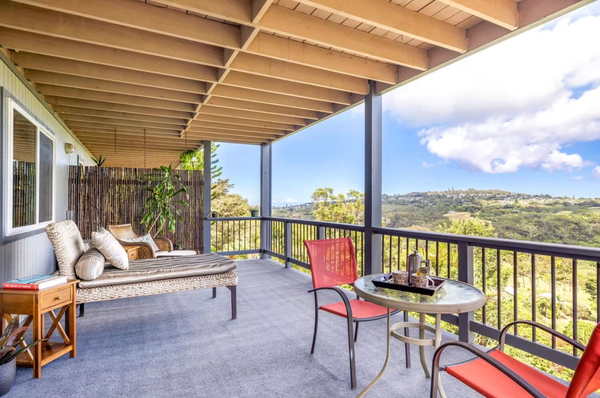 A covered balcony with a sofa, chairs, and a table, overlooking a hilly landscape.