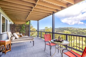 A covered balcony with a sofa, chairs, and a table, overlooking a hilly landscape.