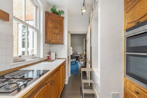 A narrow kitchen with wooden cabinets, a built-in stove, and a double oven. - GuestReady - Charming Fulham Retreat with Balcony (London)