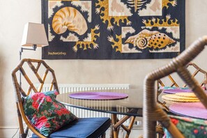 A room with a round table, wicker chairs, a radiator, and a wall hanging with abstract patterns. - GuestReady - Charming Fulham Retreat with Balcony (London)
