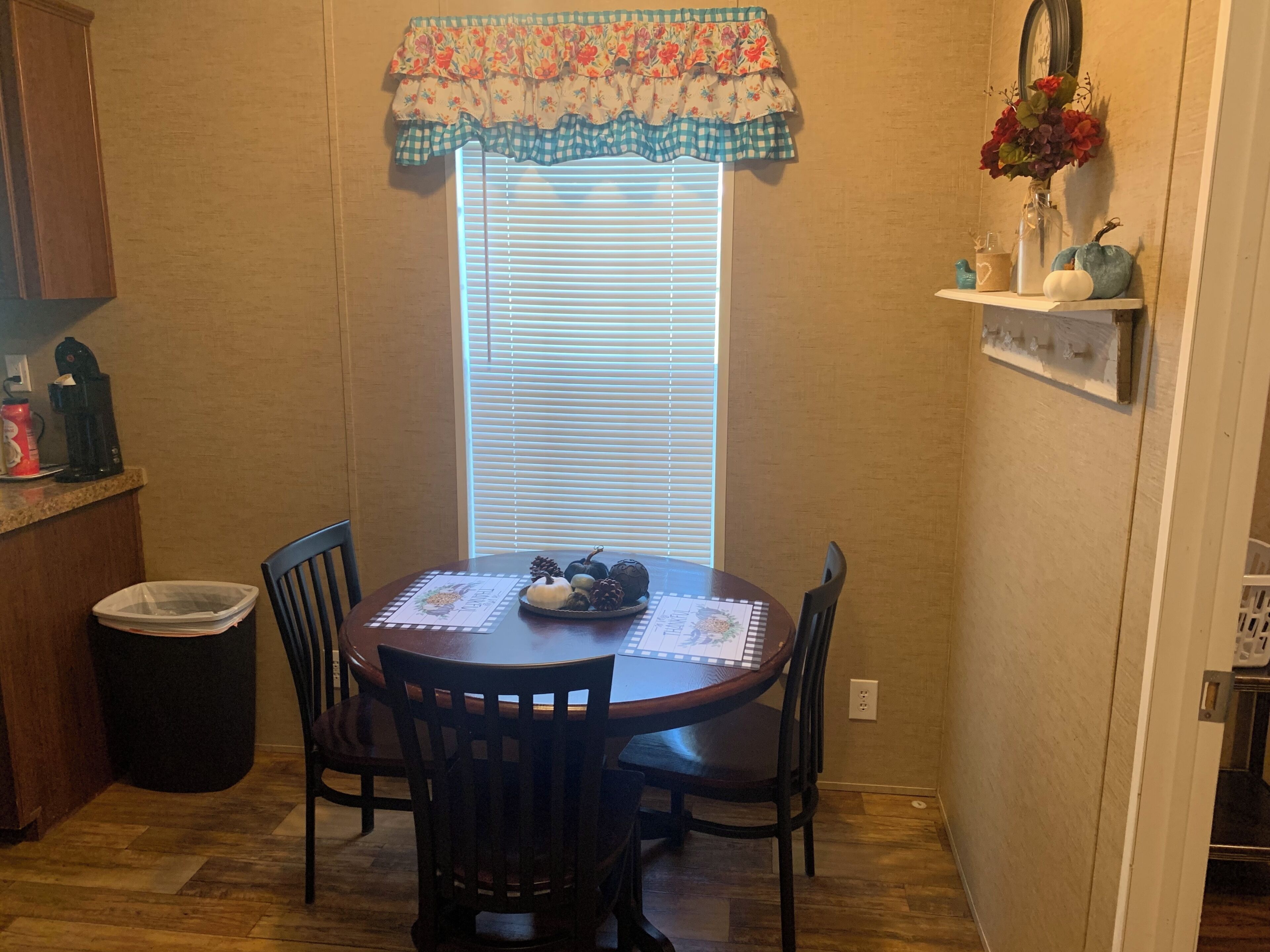 A dining area with a round table, four chairs, a window with blinds, a decorative valance, and a small shelf with a vase and pumpkins.