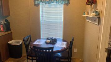 A dining area with a round table, four chairs, a window with blinds, a decorative valance, and a small shelf with a vase and pumpkins.