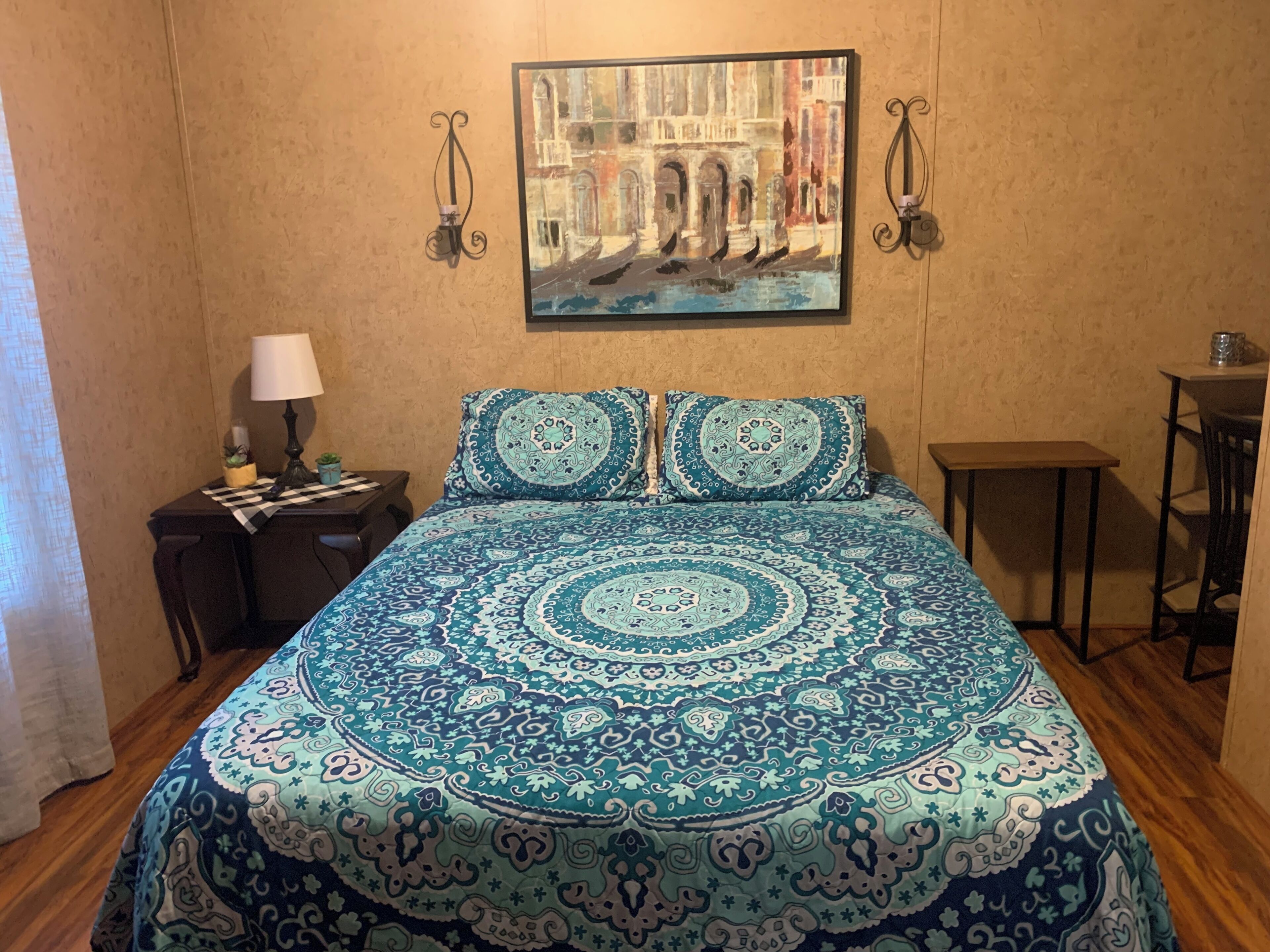 A bedroom with a patterned bedspread, a painting of a building, a nightstand with a lamp, and a small table.