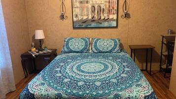 A bedroom with a patterned bedspread, a painting of a building, a nightstand with a lamp, and a small table.