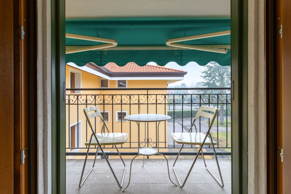 A balcony with two chairs and a small table, a metal railing, and a view of a building with a tiled roof.
