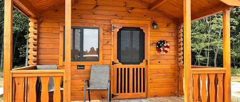 Una cabaña de madera con un porche, una puerta, una ventana, un felpudo y un adorno navideño.