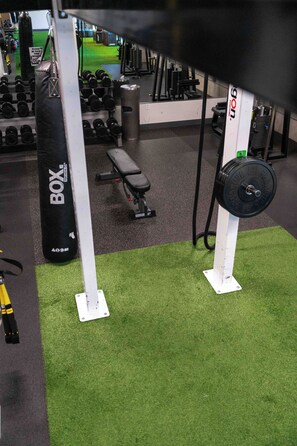 Ein Fitnessraum mit Krafttrainingsgeräten, einer Bank und einem grünen Kunstrasenboden.