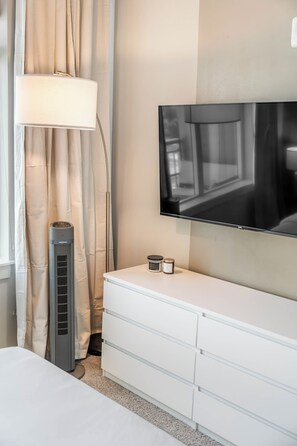 Ein Hotelzimmer mit einem wandmontierten Flachbildfernseher, einem Standheizkörper, einem Nachttisch mit Lampe und einem Vorhangfenster.