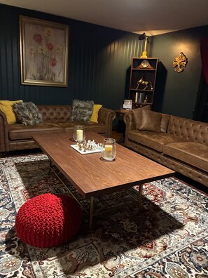 Ein Wohnzimmer mit einem braunen Sofa, einem Holztisch mit Schachfiguren und Kerzen, einem roten Pouf und einem gemusterten Teppich.