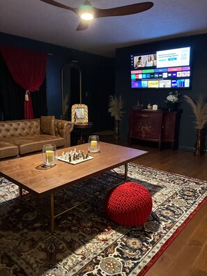 A living room with a wooden coffee table, chess set, red pouf, and a TV displaying various channels. - Luxury & Group Focused  for families or friends. 4 bedrooms 3 full baths. (Attalla)