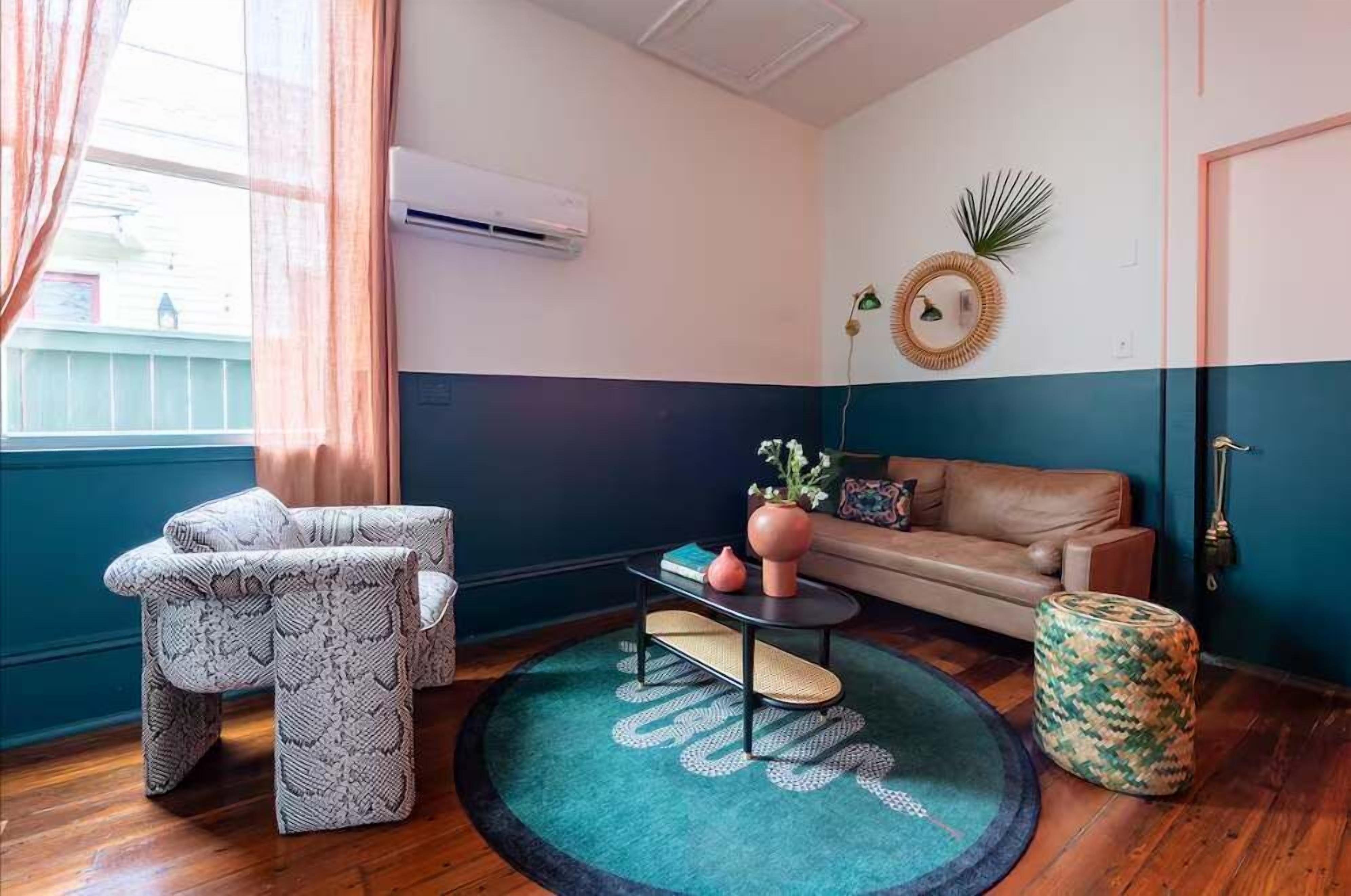 Una habitación con un sofá, un sillón estampado, una mesita con un jarrón, un tapete redondo y un aire acondicionado instalado en la pared.