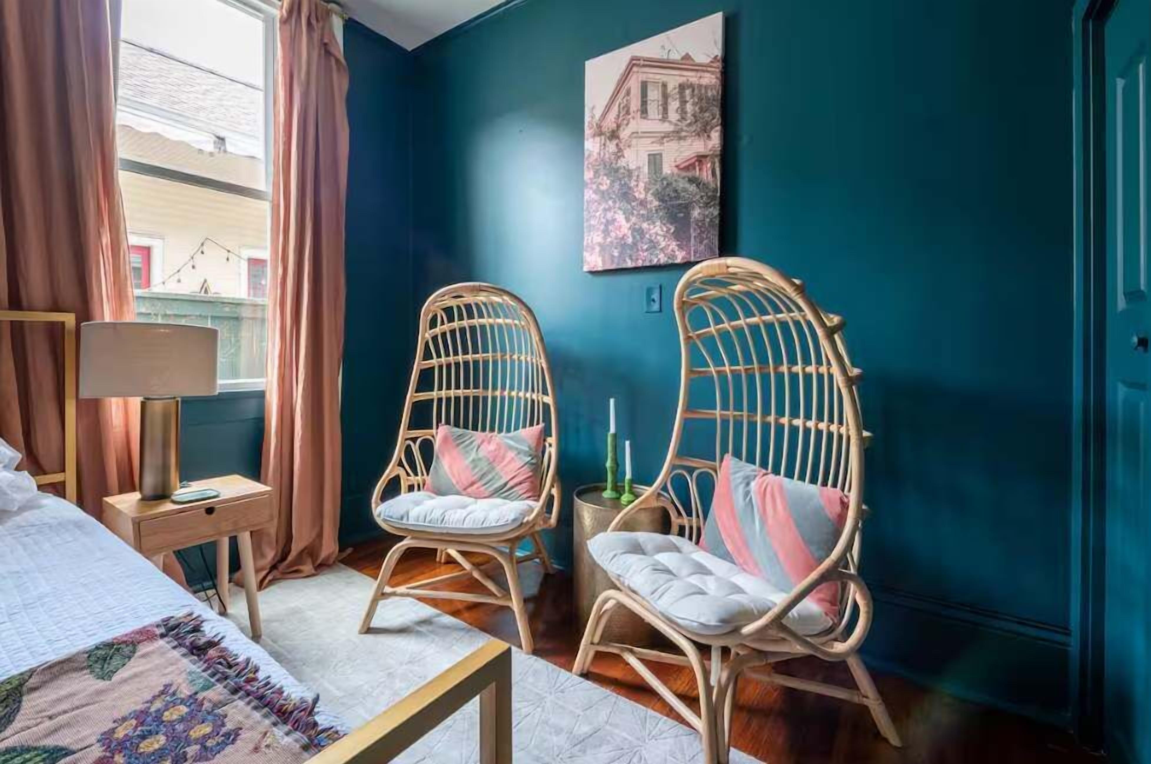 A room with a bed, two wicker chairs, a side table with a lamp, and a painting on the wall.