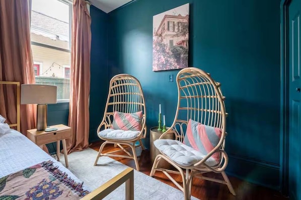 Une pièce comprenant un lit, deux fauteuils en osier, une table d’appoint avec une lampe et un tableau accroché au mur.