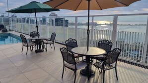 A rooftop pool area with a table and chairs, a green umbrella, and a view of the city skyline and water.