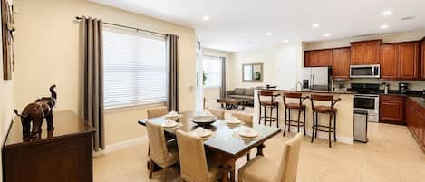 A spacious living area with a dining table, kitchen, and a large window with curtains.