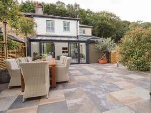 A patio area with a dining table and chairs, a glass-enclosed structure, and a garden with potted plants. - Cottage by the River (Burnley)